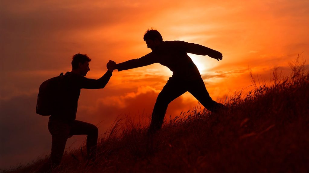 Mann reicht helfend beim Bergsteigen die Hand eines anderen Mannes im Sonnenuntergang.